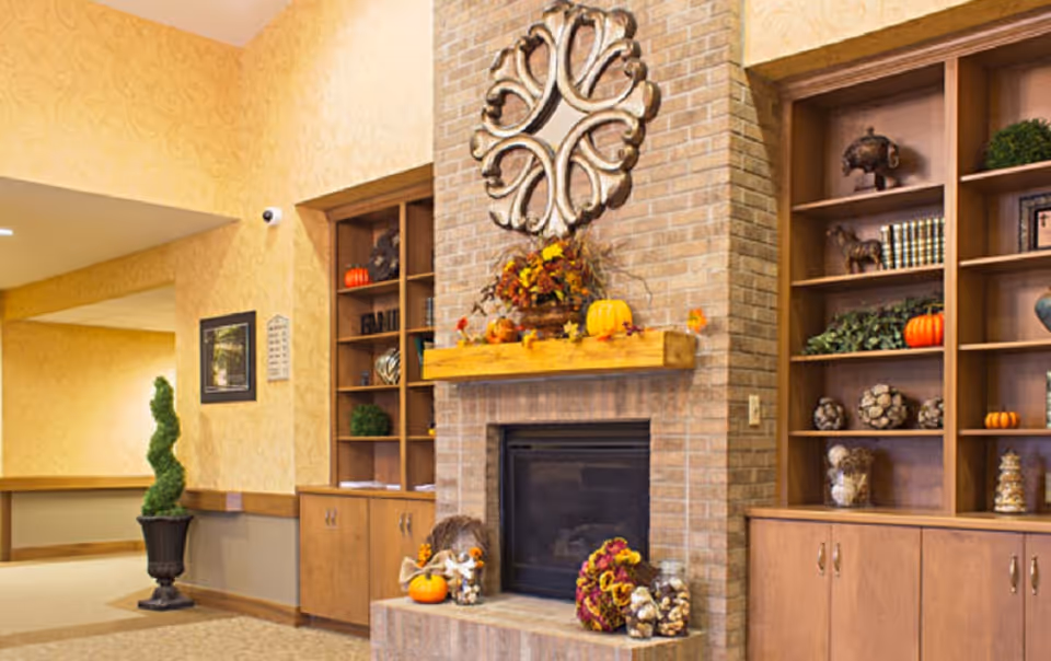 A cozy interior common area featuring a brick fireplace decorated with autumn-themed items such as pumpkins and floral arrangements. On either side of the fireplace are wooden built-in shelves and cabinets displaying decorative objects and greenery. The walls are painted in warm tones with a patterned wallpaper, and a topiary plant is placed in a black pot near the hallway entrance.