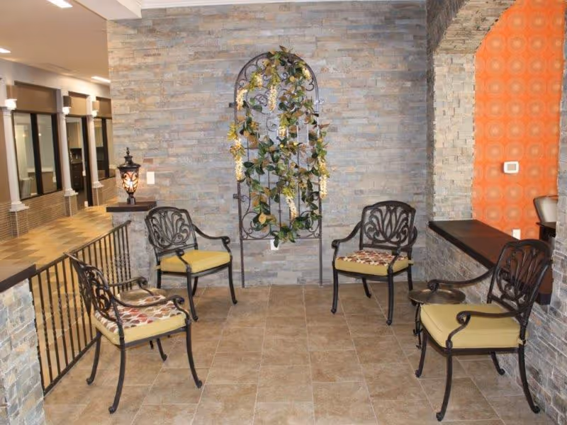 A cozy indoor seating area with four metal chairs featuring patterned cushions arranged around a small table. The space has stone tile flooring and stone walls, with a decorative metal trellis adorned with green and yellow foliage mounted on the wall. There is a warm lamp on a stone ledge and an orange patterned accent wall on the right side.
