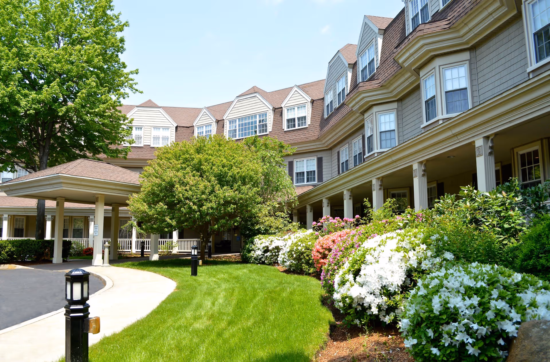 Exterior view of Grove Manor Estates showing a multi-story building with beige siding and multiple windows. The building is surrounded by well-maintained green lawns, colorful flowering bushes, and trees. A covered entrance with columns is visible on the left side, along with a paved driveway and walkway.