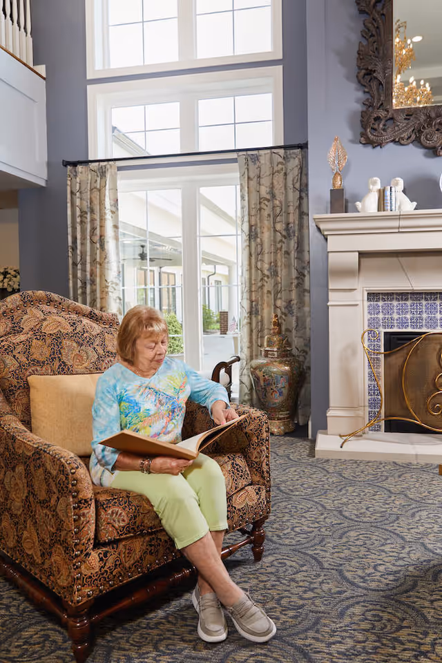 An elderly woman sitting on a patterned armchair in a cozy living room, reading a book. The room features large windows with floral curtains, a decorative fireplace with blue and white tiles, a large ornate mirror above the mantel, and a patterned carpet.