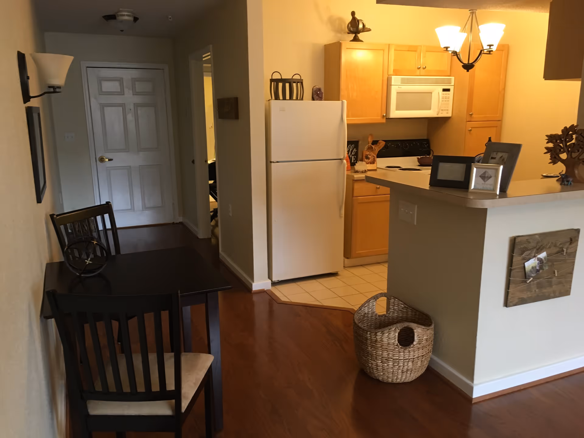 Interior view of a senior living facility apartment showing a small dining area with a dark wooden table and two chairs, a kitchen with light wood cabinets, a white refrigerator, a microwave above a stove, and a countertop with framed photos and decorative items. There is a woven basket on the floor near the counter and a hallway leading to a closed door.