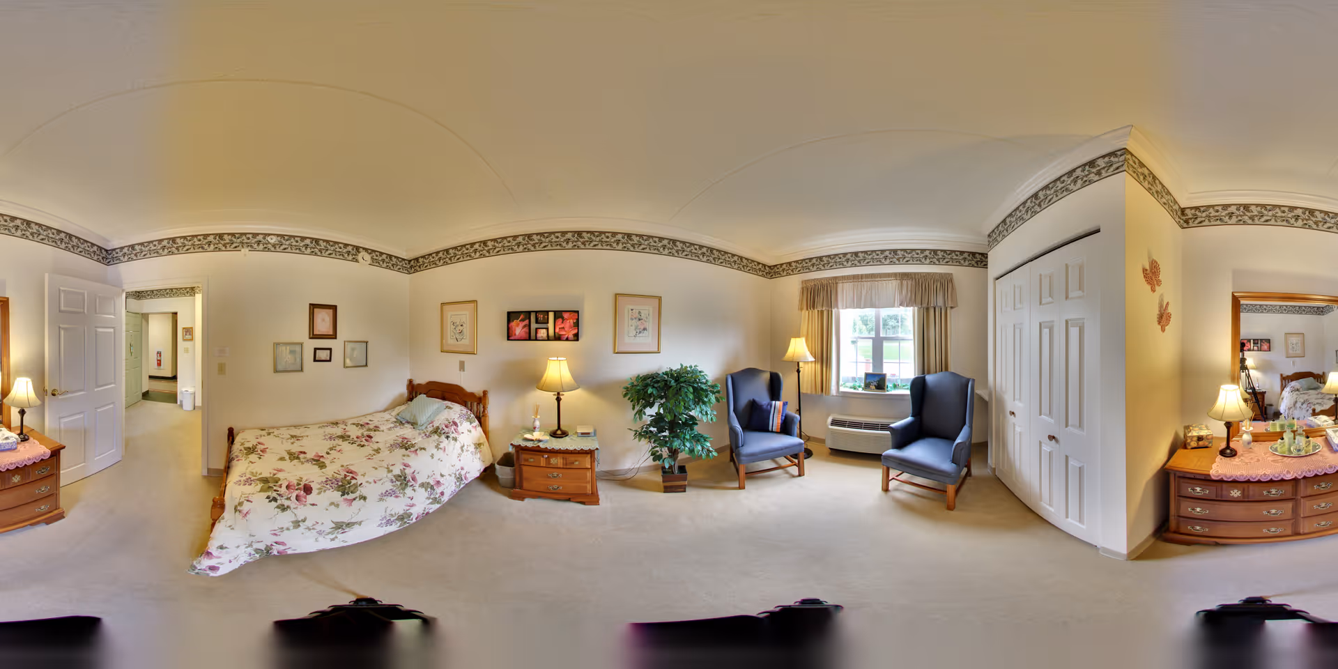 Bright traditional bedroom with a floral-covered bed, two nightstands with lamps, two blue armchairs by a window, a dresser and a closet.