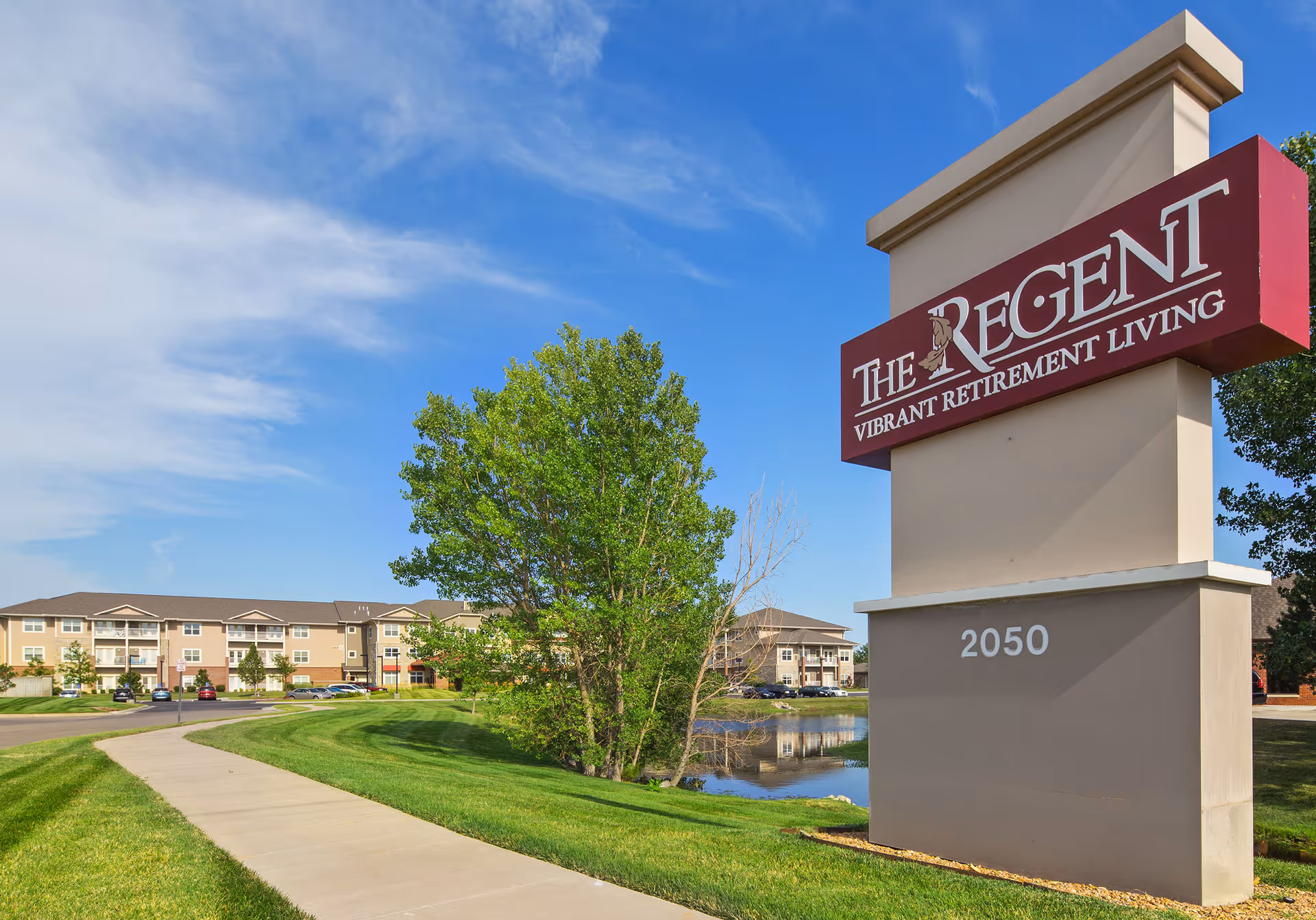 Large entrance sign reading 'The Regent Vibrant Retirement Living' beside a sidewalk and lawn with the apartment building and pond in the background.