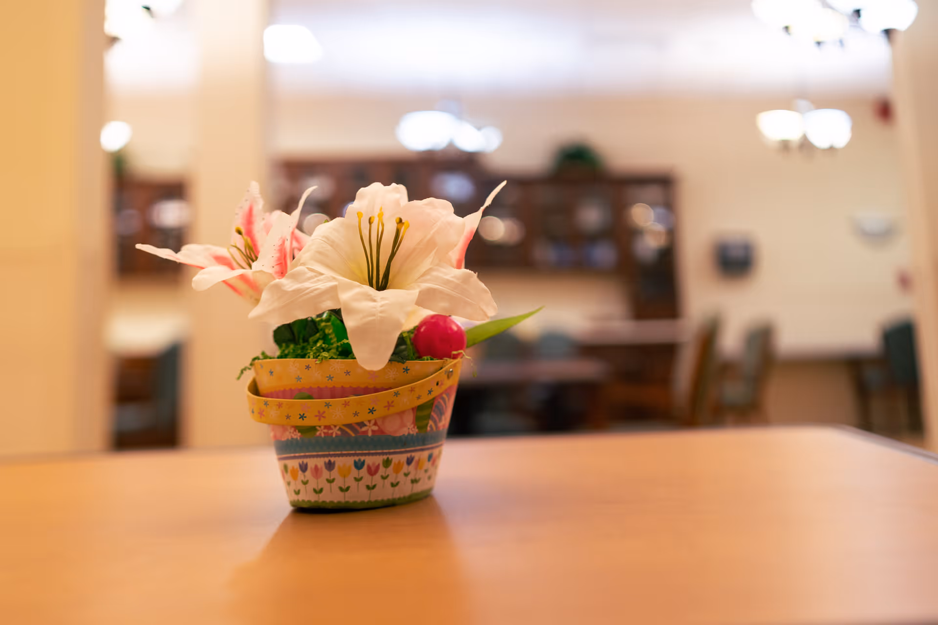 A small decorative flower arrangement with white and pink flowers in a colorful pot placed on a wooden table in a softly lit room with blurred background showing chairs and cabinets.