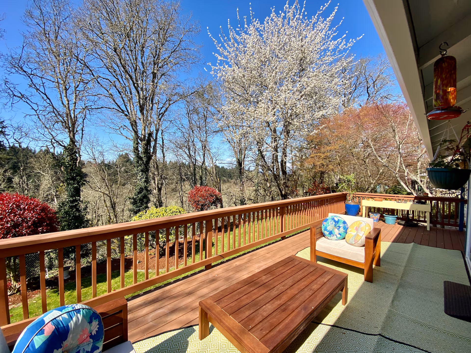 A sunny outdoor wooden deck with a wooden coffee table and cushioned chairs with colorful floral pillows. The deck overlooks a garden with various trees, including one with white blossoms, and shrubs with red and green foliage. The sky is clear and blue.