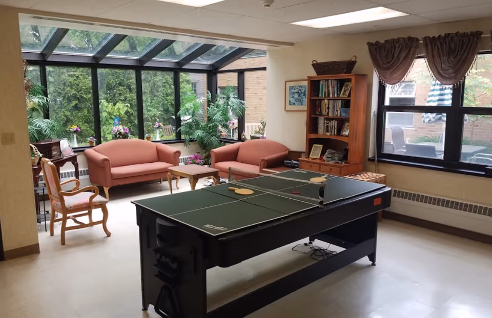 Sunlit common room with a ping-pong table in the center, couches and chairs by large windows and a bookshelf.