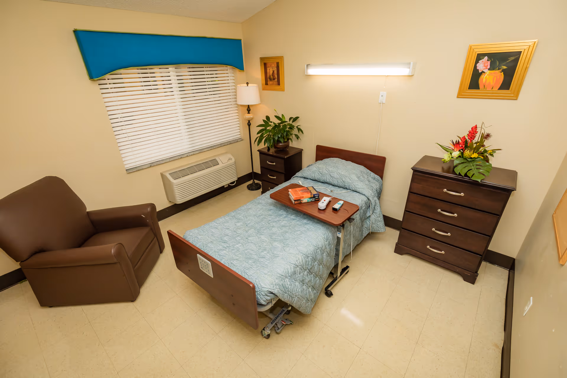 A tidy single-occupancy rehab bedroom with a hospital bed, overbed tray holding remotes and books, a dresser, armchair, lamp, and a window with blinds.