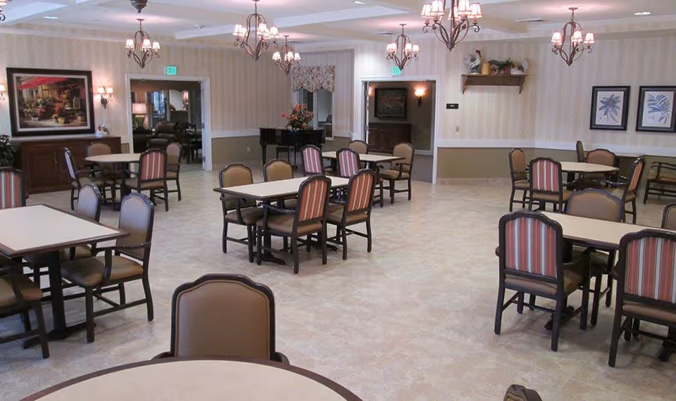 A spacious dining room with multiple tables and chairs arranged neatly. The room features beige tiled flooring, cream-colored walls with a chair rail, and several chandeliers hanging from the ceiling. There are framed pictures on the walls, a piano in the back corner, and floral arrangements on a table near the doorway.
