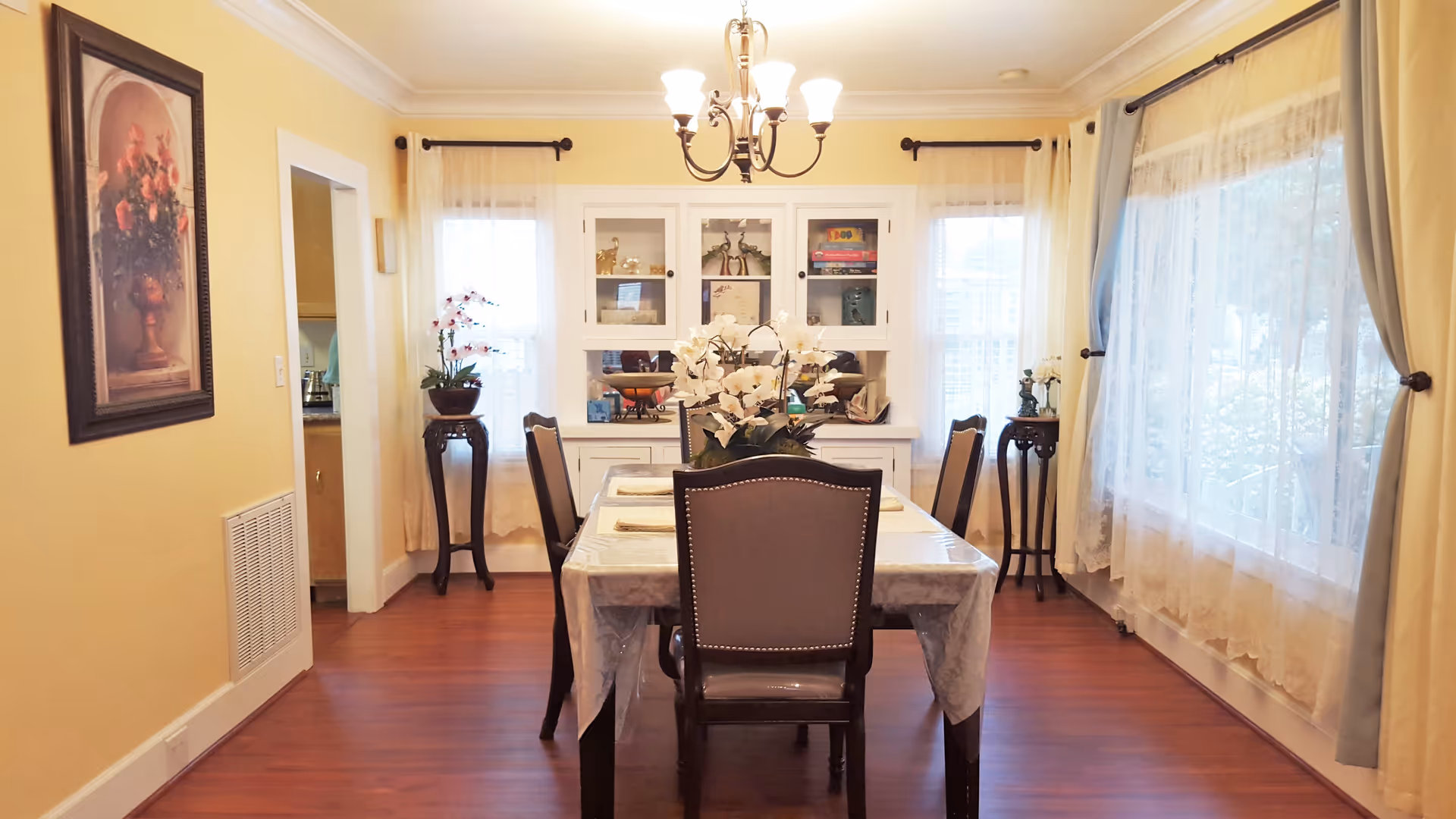 A cozy dining room with a rectangular table covered with a plastic tablecloth and set with placemats. Six upholstered chairs surround the table. A chandelier with six lights hangs above. The room has yellow walls, wooden floors, and large windows with sheer curtains letting in natural light. There is a white cabinet with glass doors at the far end of the room, displaying decorative items and board games. Two small tables with potted plants flank the cabinet, and a framed floral painting hangs on the left wall.