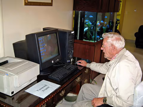 An elderly man sitting at a wooden desk using a desktop computer with a CRT monitor. There is a printer on the desk and a sign that reads 'Password Assist'. Behind the desk is a large fish tank with colorful fish. The setting appears to be a common area or lounge.