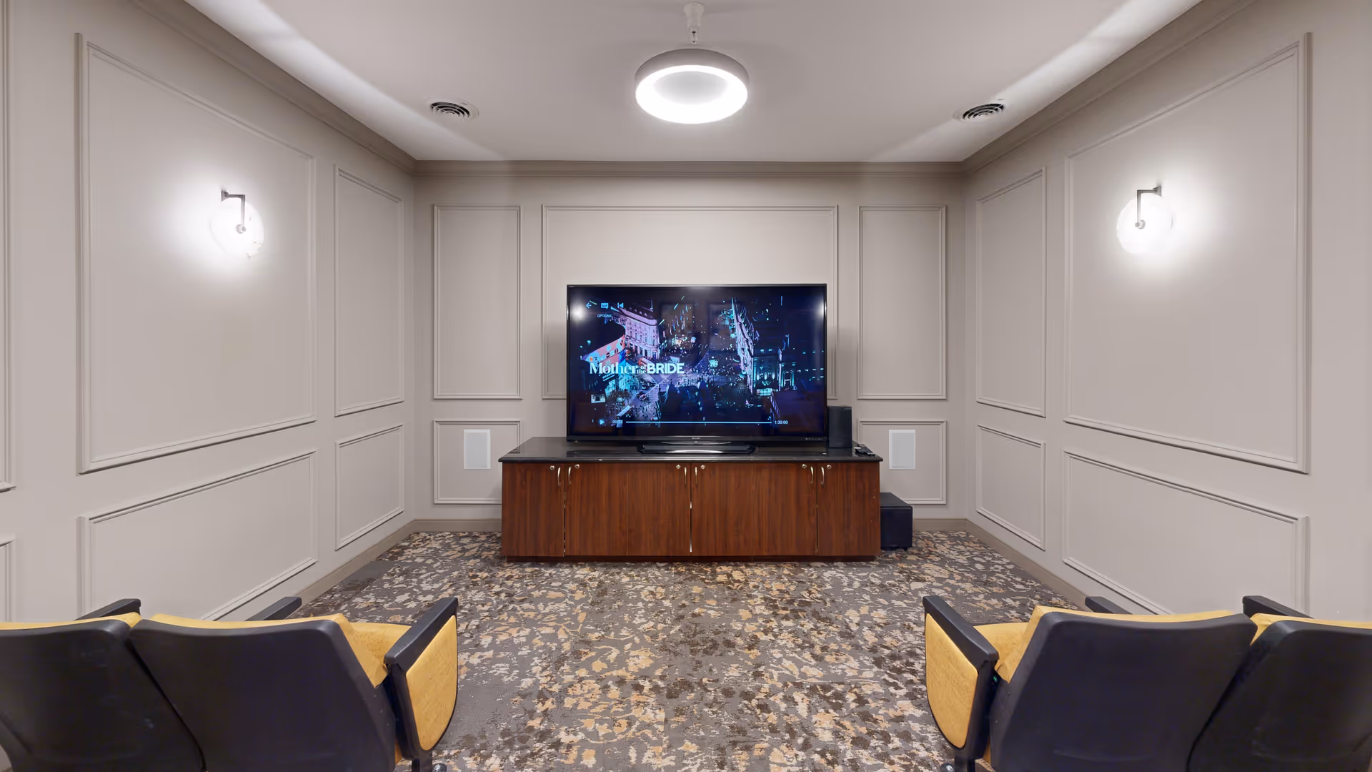 A small media room with a large flat-screen TV mounted on a wooden cabinet. The TV is playing a movie titled 'Mother BRIDE'. The room has patterned carpet flooring, light gray paneled walls, two wall-mounted lights, and several theater-style chairs with black backs and yellow seats facing the TV.