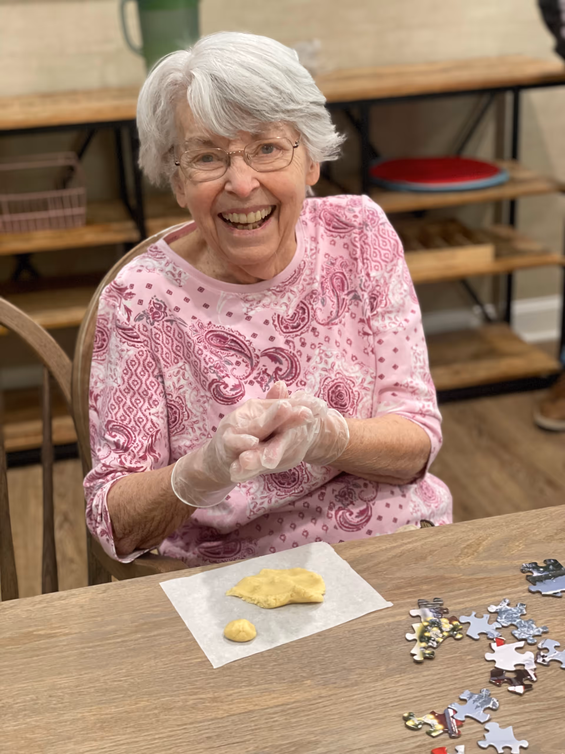 An elderly woman with white hair and glasses, wearing a pink patterned shirt and disposable gloves, is sitting at a wooden table. She is smiling and appears to be shaping dough on a piece of parchment paper. There are puzzle pieces scattered on the table to her right. In the background, there is a wooden shelf with various items on it.