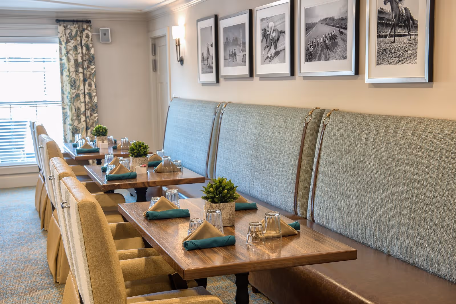 A dining area with wooden tables set with folded beige and teal napkins, upside-down drinking glasses, and small potted plants. The seating includes cushioned chairs and a long upholstered bench against the wall. Above the bench are five black and white framed photographs depicting horse racing scenes. A window with floral curtains allows natural light into the room.