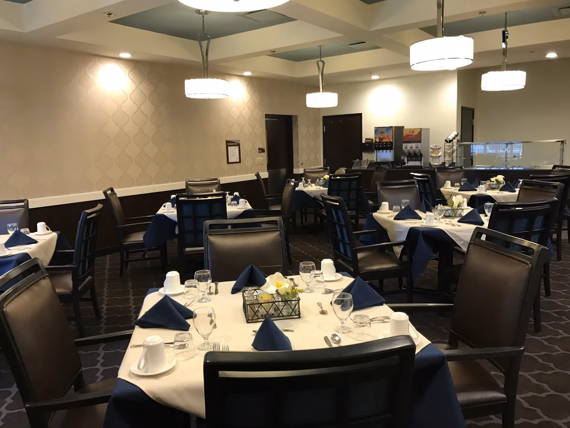 Well-lit dining room with multiple tables set with white tablecloths, blue napkins, glassware, and chairs.