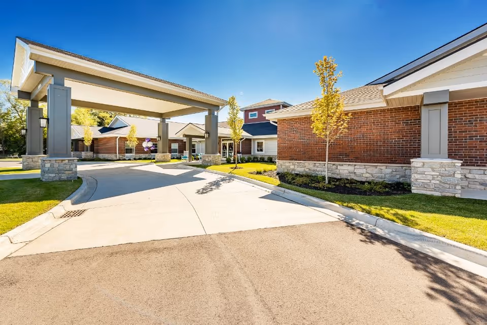 Exterior view of Provision Living at Livonia showing a covered driveway entrance with brick and stone buildings, small trees, and a clear blue sky.