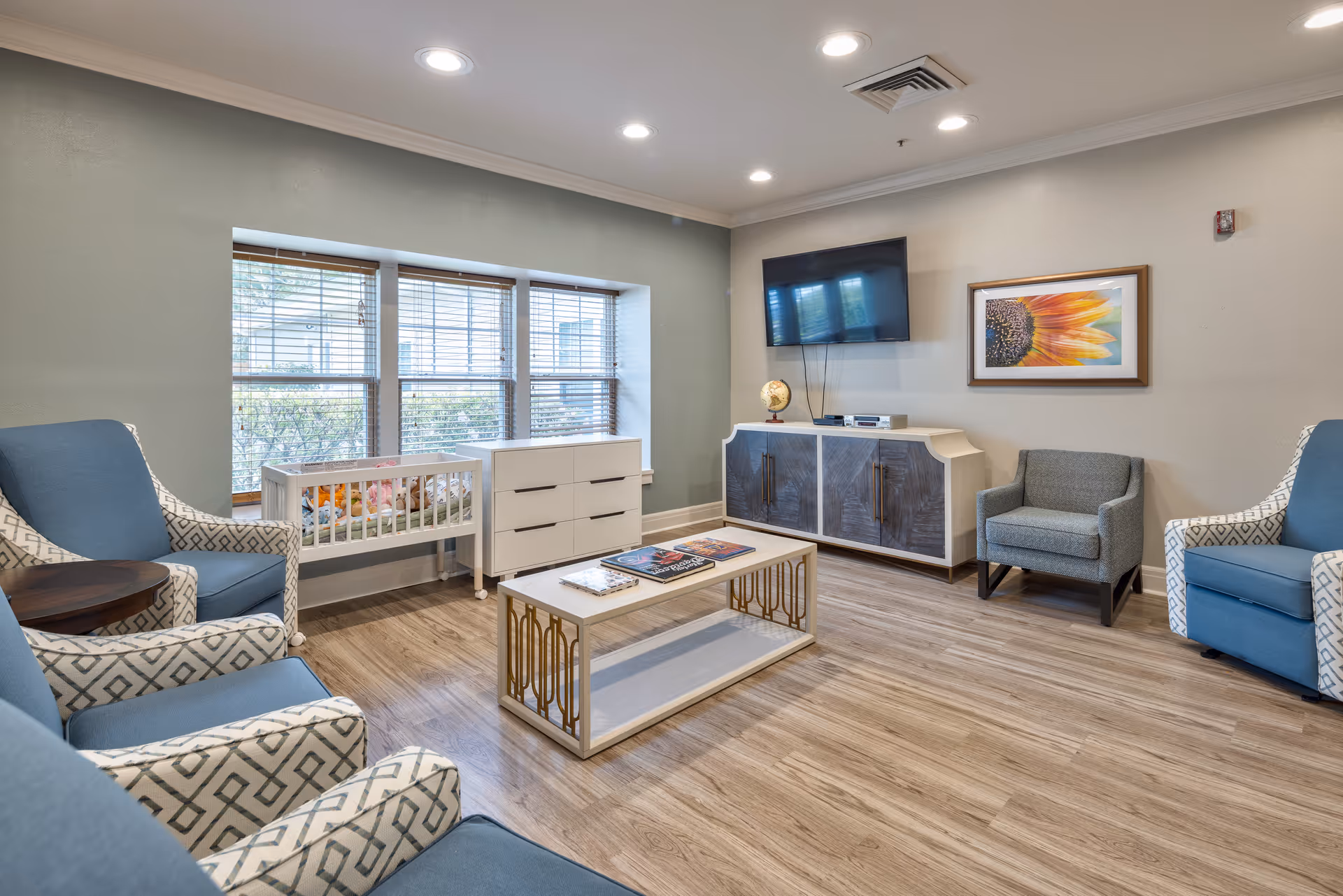 A bright and cozy living room with light wood flooring, blue and patterned armchairs arranged around a white coffee table with gold accents. A white dresser and a small crib with stuffed animals are positioned under a set of three windows with blinds. On the opposite wall, a flat-screen TV is mounted above a modern cabinet, next to a framed sunflower artwork. The ceiling has recessed lighting.