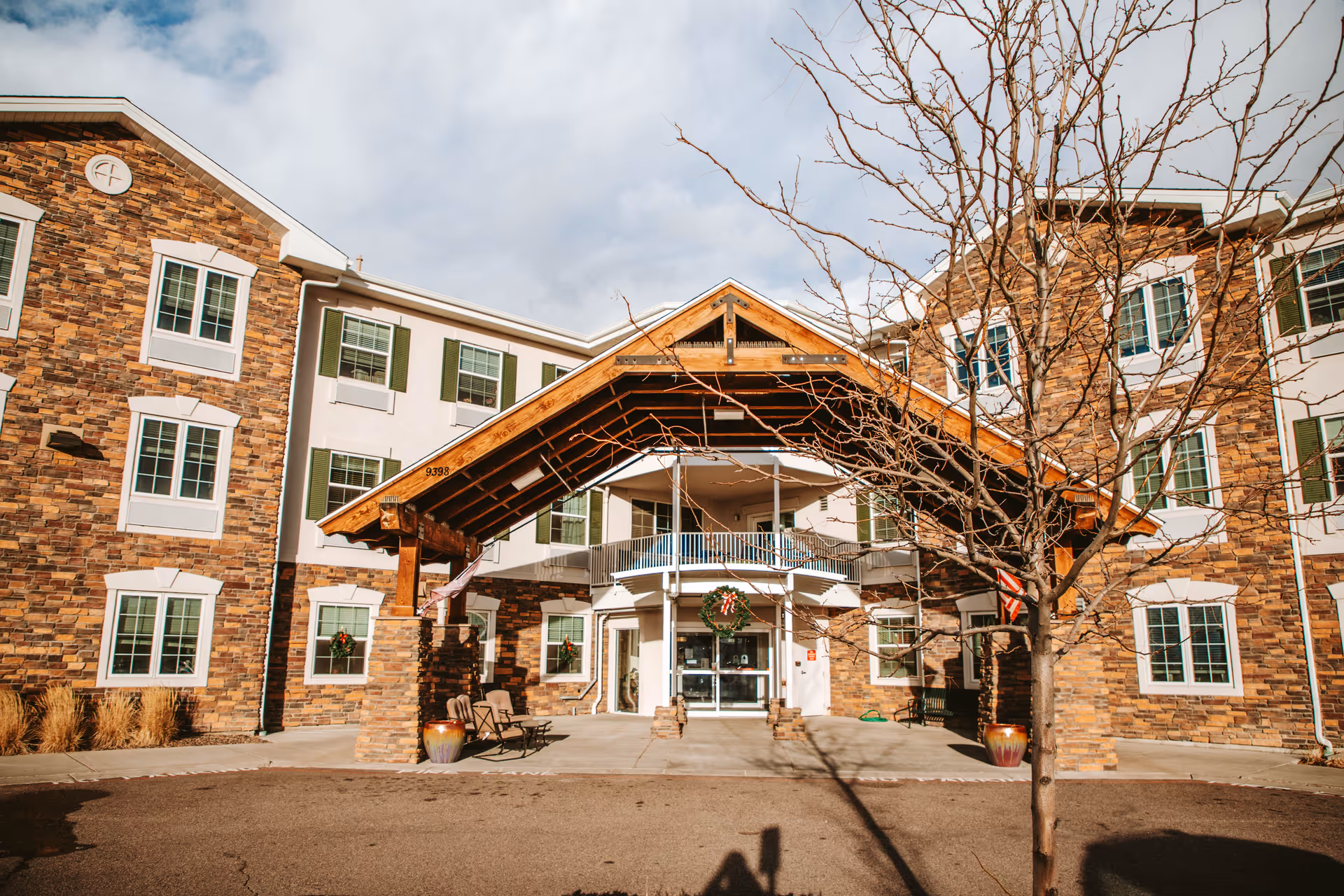 Exterior front view of The Suites Parker senior living facility showing a three-story building with stone and white siding, multiple windows with green shutters, a covered entrance with wooden beams, and a leafless tree in the foreground.