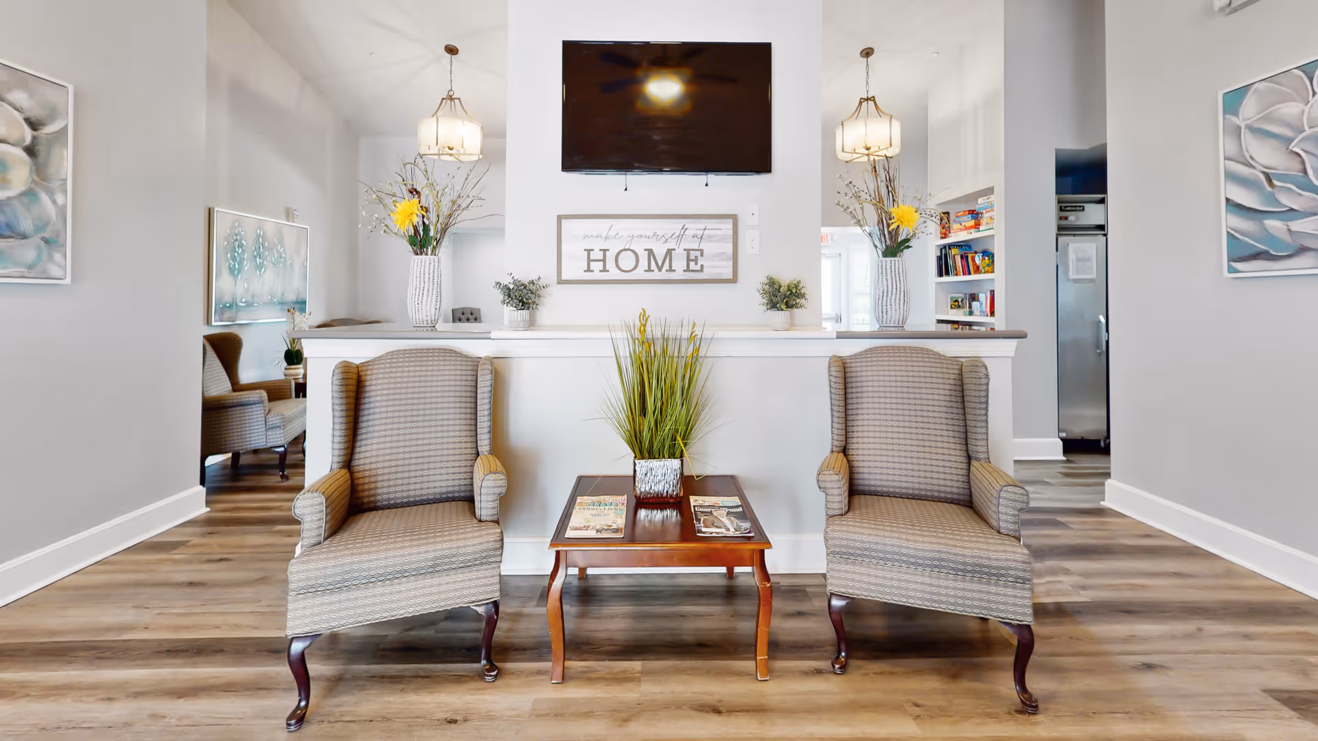 A cozy seating area in a senior living facility with two upholstered armchairs facing a wooden coffee table that holds magazines and a potted plant. Behind the chairs is a white half-wall with decorative vases containing flowers and branches. Above the wall is a mounted flat-screen TV and a sign that reads 'make yourself at HOME'. The room has light-colored walls, wood flooring, and framed floral artwork on the walls.
