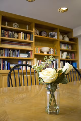 A wooden table with a glass jar holding white roses and small green flowers in the foreground. Behind the table are black wooden chairs and a large wooden bookshelf filled with books and decorative items. The ceiling has recessed lighting.