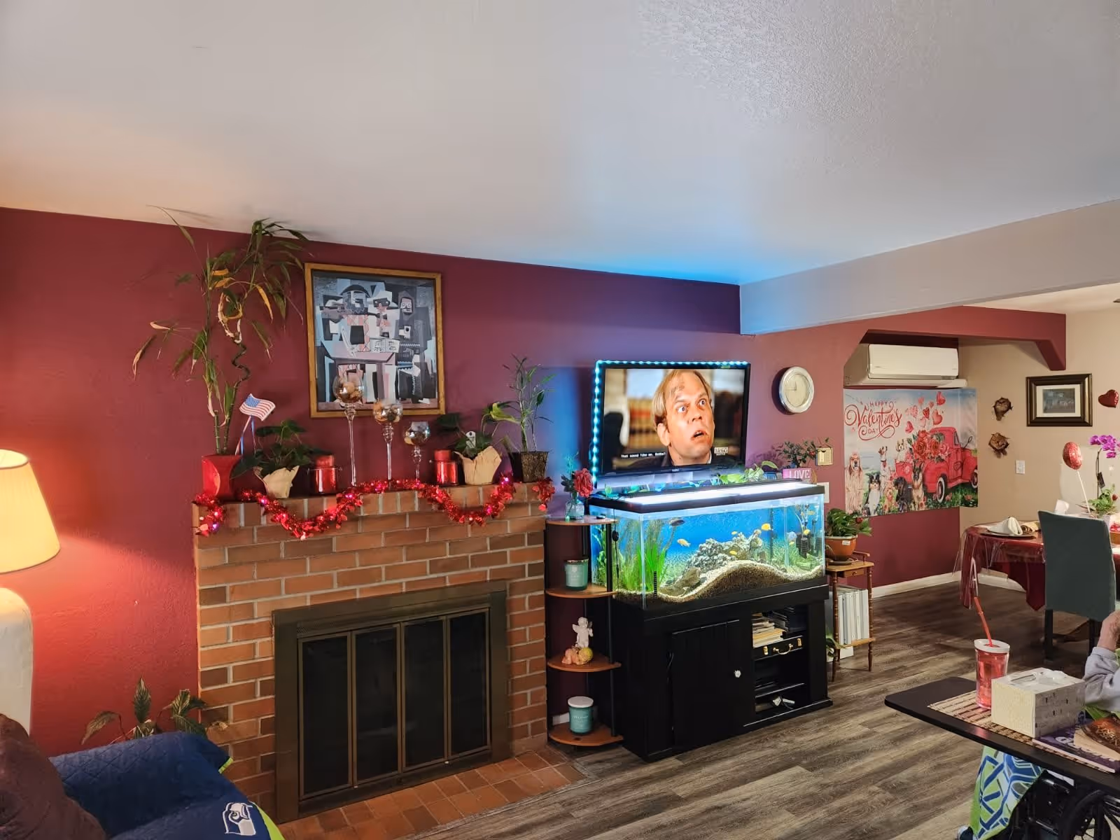 A cozy living room with a brick fireplace decorated with plants, candles, and red garland. Above the fireplace is a framed abstract painting. To the right, there is a large fish tank with colorful fish, and above it, a TV screen displaying a surprised man. The walls are painted burgundy and beige, and there is a Valentine's Day poster on the far wall. A dining area with a table and chairs is partially visible in the background.