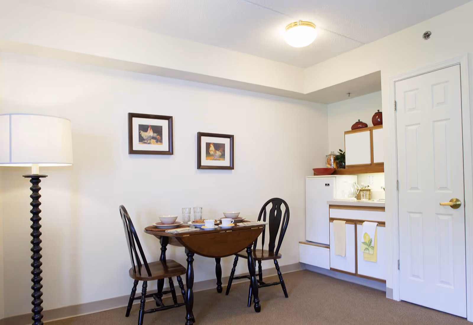 Small dining area with a round wooden table set for two, two chairs, a tall floor lamp, and a kitchenette in the background.
