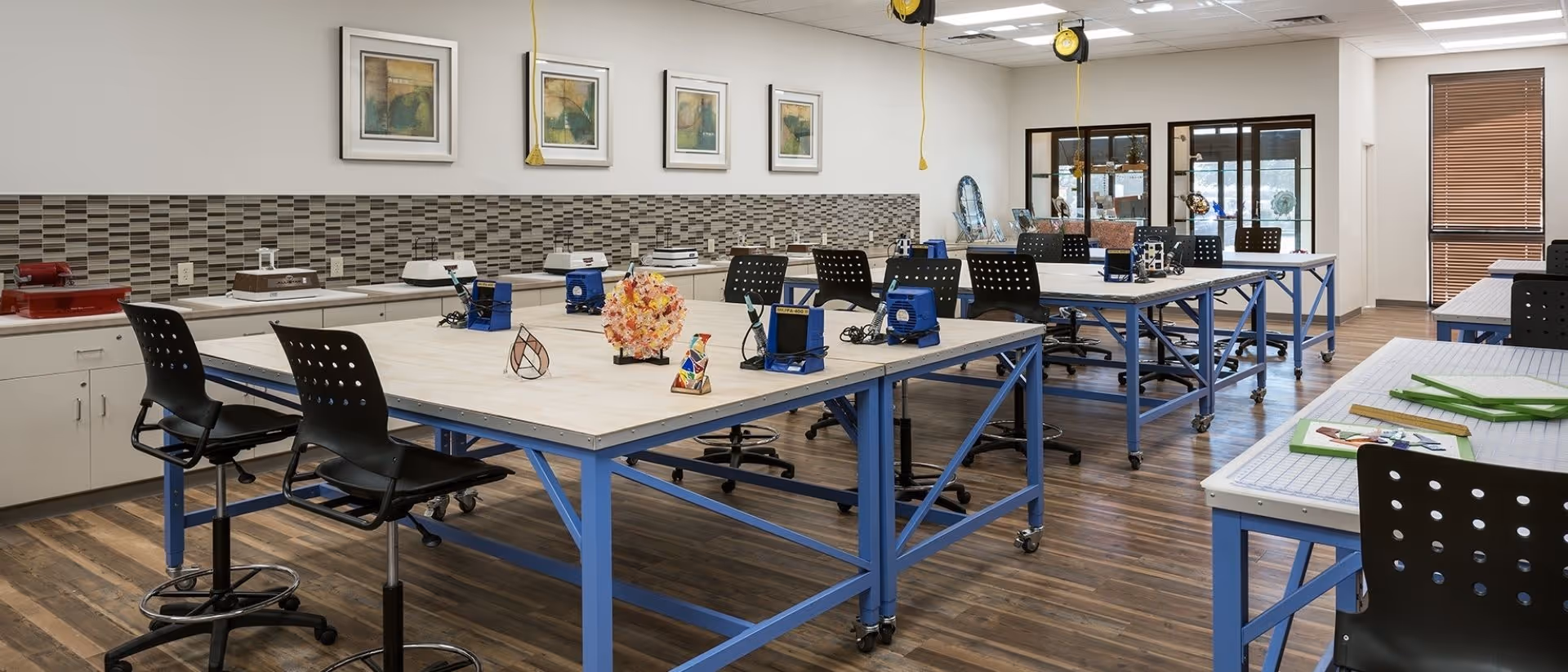 A spacious, well-lit craft or workshop room with multiple large tables on blue metal frames, black swivel chairs, and various craft tools and materials placed on the tables. The room has wooden flooring, a tiled backsplash along one wall, and framed artwork hanging above the backsplash. Large windows with blinds allow natural light to enter the space.