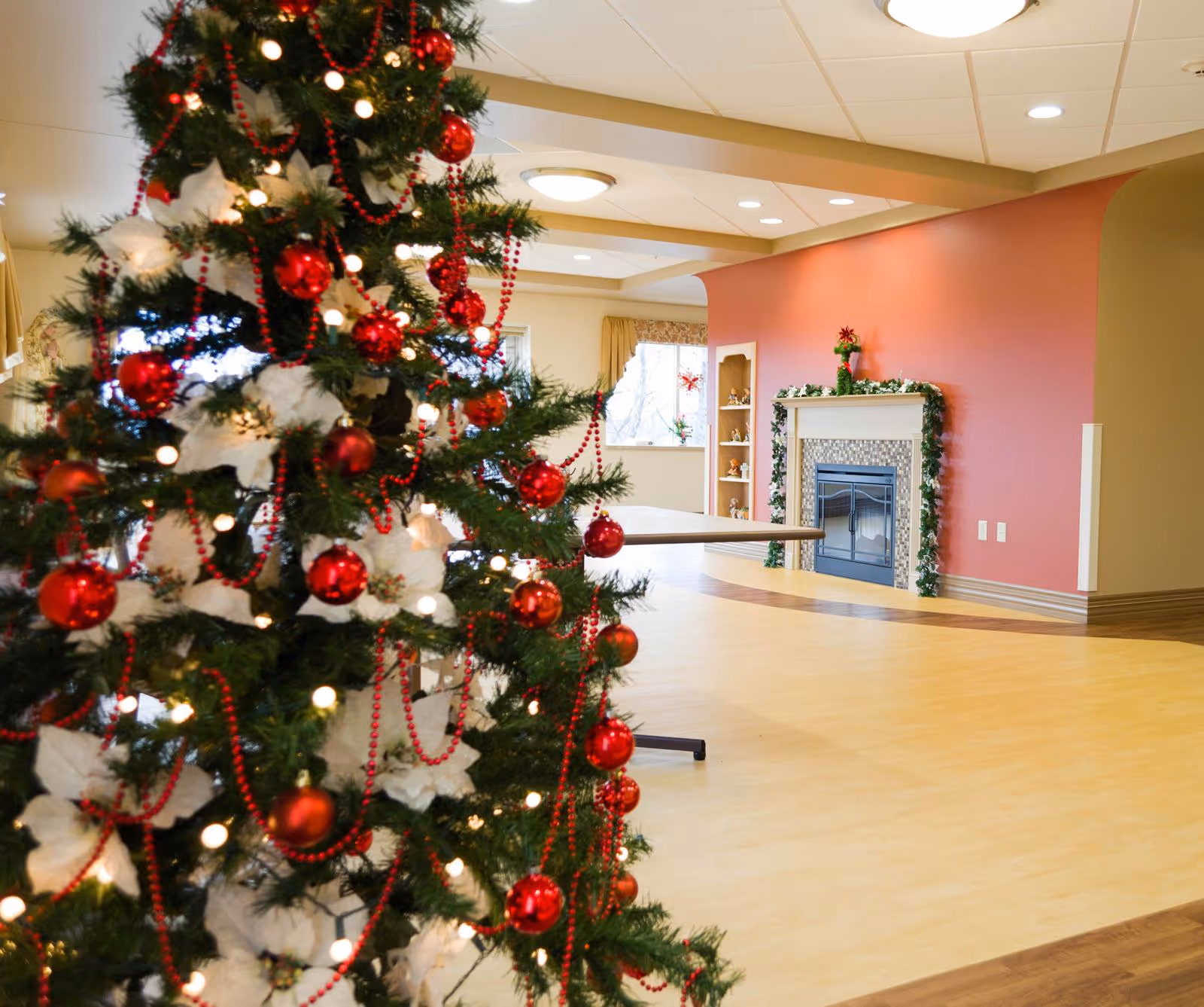 A decorated Christmas tree with red ornaments and white flowers stands in the foreground of a spacious room with light wood flooring. In the background, there is a fireplace adorned with garland and a red wall, along with a window and built-in shelves with small decorations.
