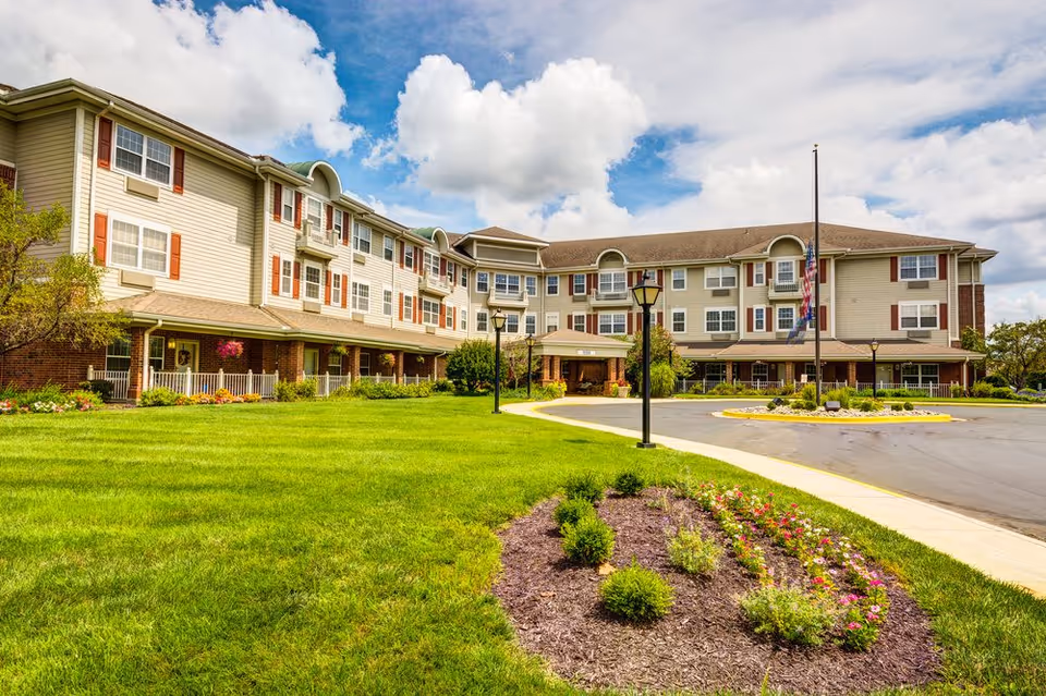 Exterior view of a three-story senior living facility building with beige siding and red shutters. The building is surrounded by a well-maintained green lawn, flower beds, and a paved driveway with lamp posts. The sky is partly cloudy.