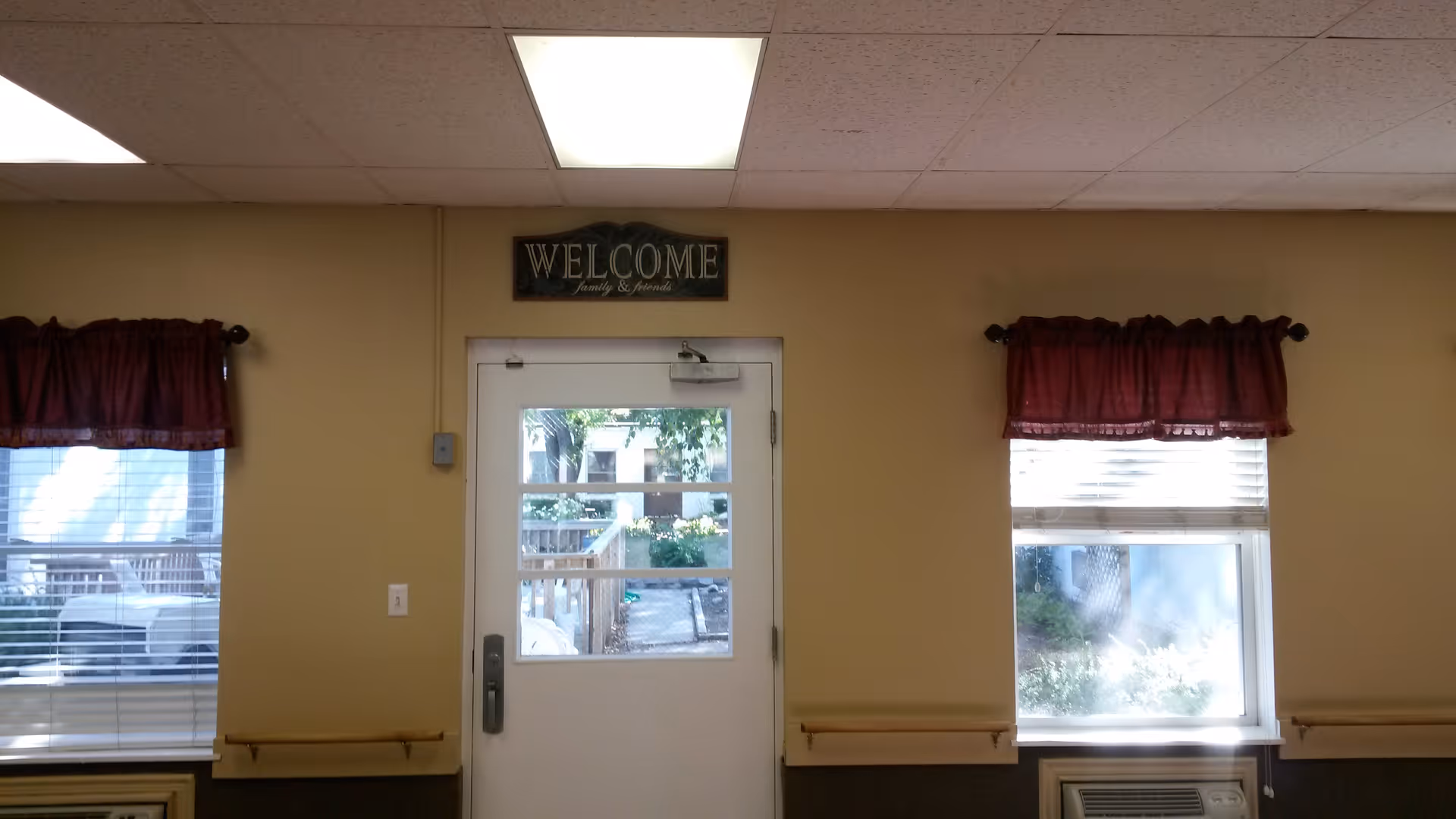 Interior view of a room with beige walls featuring a white door with glass panels in the center. Above the door is a sign that reads 'WELCOME Family & Friends'. On either side of the door are two windows with maroon valances and white blinds. Outside the windows, some greenery and outdoor structures are visible. The ceiling has white tiles with a fluorescent light panel.