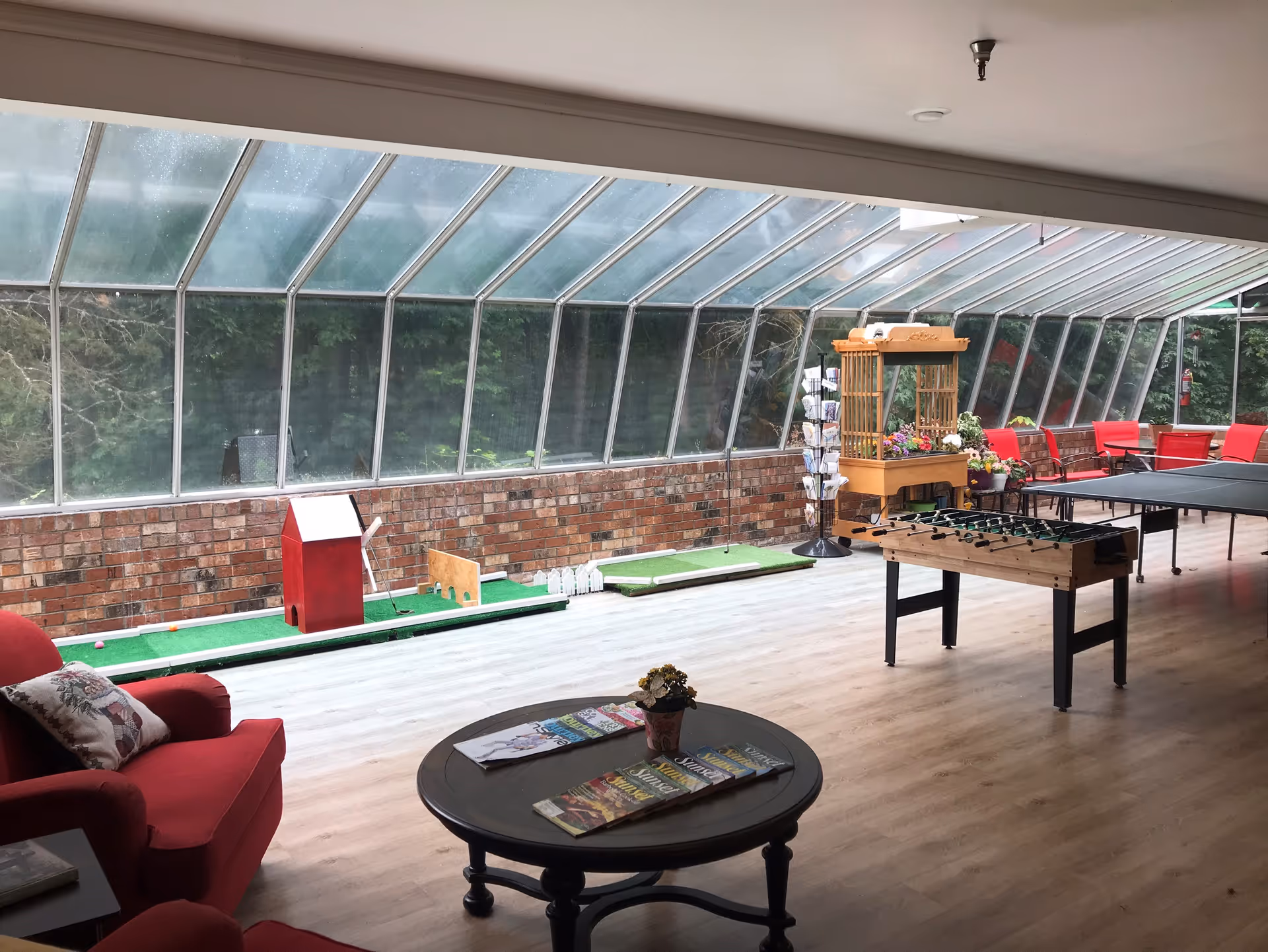 A bright indoor recreational room with large slanted glass windows along one side, featuring a mini putting green, a foosball table, a ping pong table, red cushioned chairs, a round wooden table with magazines and a small flower pot, and a wooden display stand with flowers and cards.