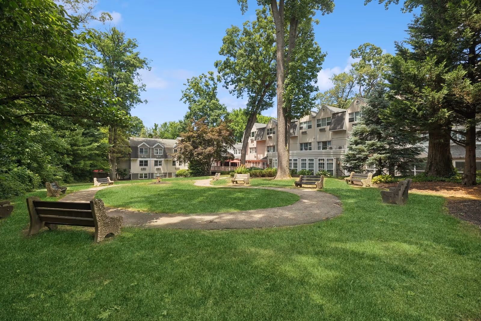 Outdoor view of a senior living facility with a circular walking path surrounded by green grass and several benches. The building is visible in the background with multiple windows and surrounded by tall trees under a clear blue sky.