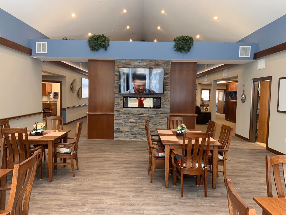Communal dining area with wooden tables and chairs facing a stone fireplace wall and a wall-mounted TV in a senior living facility.