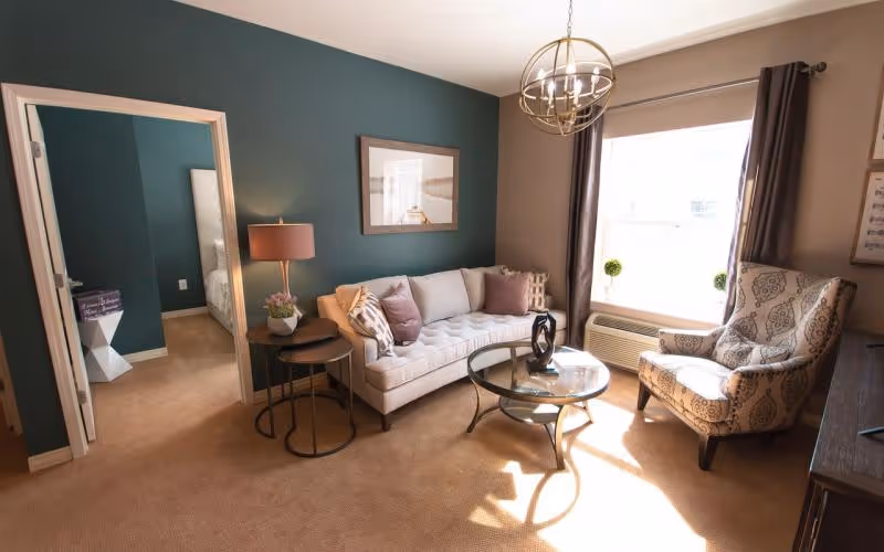 Sunlit living room featuring a tufted sofa, patterned armchair, round glass coffee table, teal accent wall and a doorway to a bedroom.