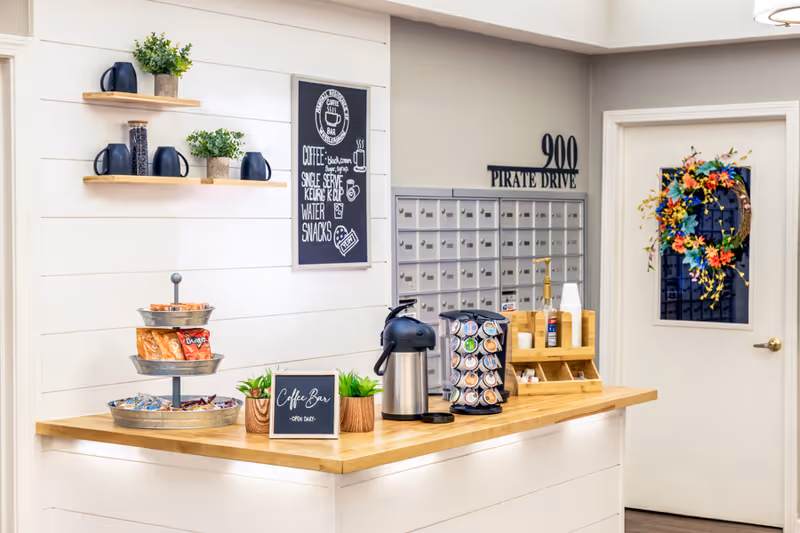 Bright interior mailroom and coffee bar with a counter holding a coffee station and snacks, wall mailboxes, and a door with a floral wreath.