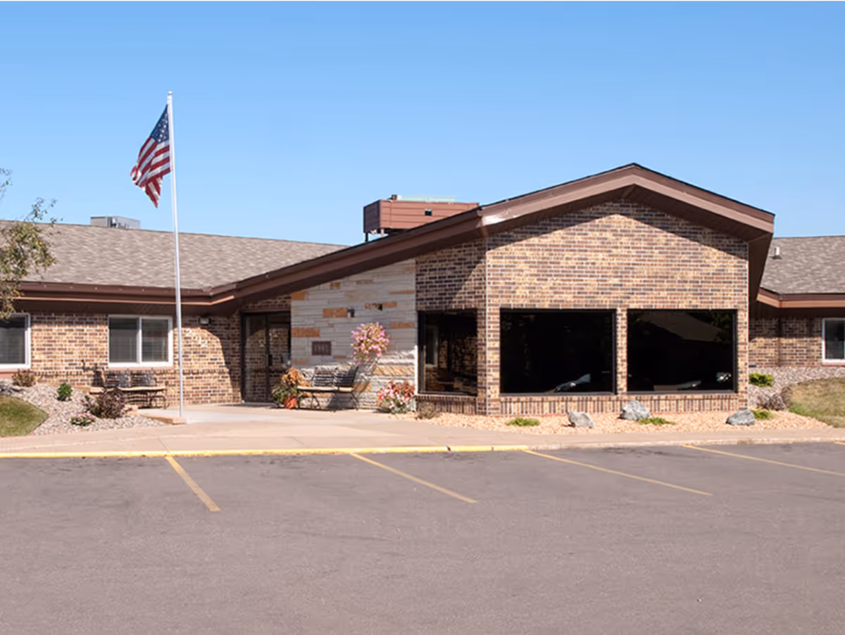 Brick single-story senior living facility entrance with large windows, an American flag, benches, and a parking area in front.