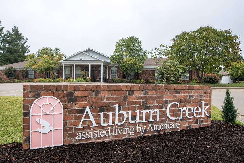 Brick sign for Auburn Creek assisted living by Americare in front of a single-story brick building with white columns and a small porch, surrounded by trees and greenery.