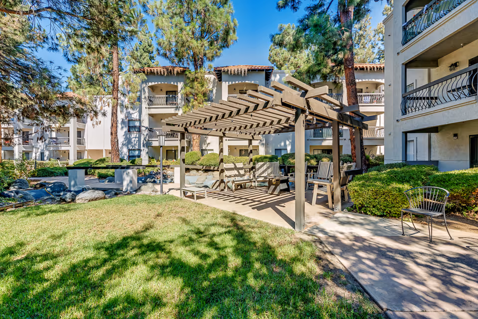 Outdoor courtyard area of a senior living facility with a wooden pergola providing shade over several chairs and benches. The surrounding area features green grass, bushes, tall pine trees, and a multi-story building with balconies in the background under a clear blue sky.