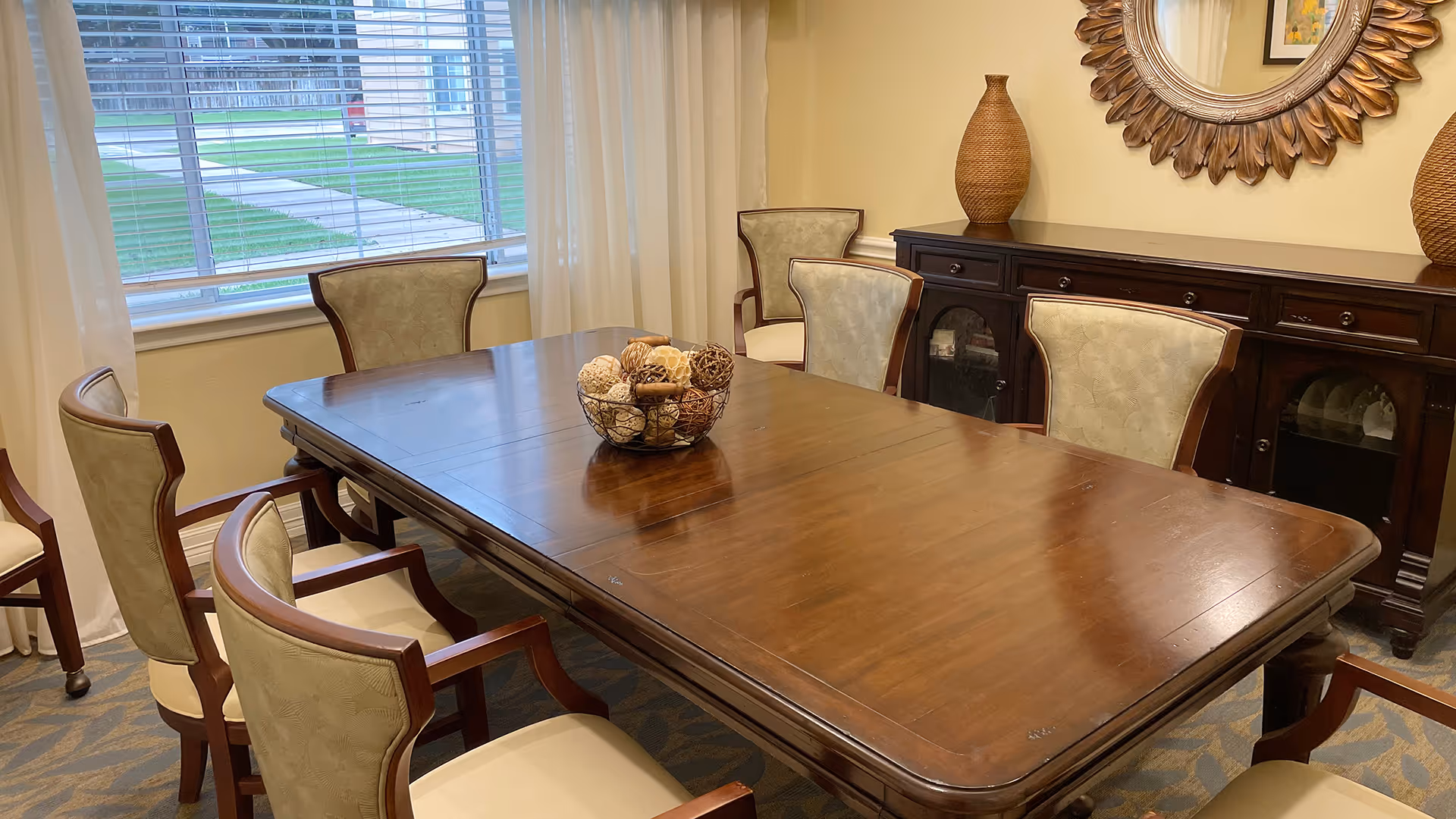 A dining room with a large wooden table surrounded by six upholstered chairs. On the table is a decorative bowl filled with assorted natural elements. Behind the table is a dark wooden sideboard with two large woven vases and a round decorative mirror hanging above it. A window with white blinds and curtains lets in natural light.