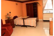 A senior living facility bedroom with an orange wall, a single bed with a patterned bedspread, a red armchair, a wooden nightstand with a flower arrangement, a wooden wardrobe, a window with floral curtains, and a small table on wheels positioned over the bed.