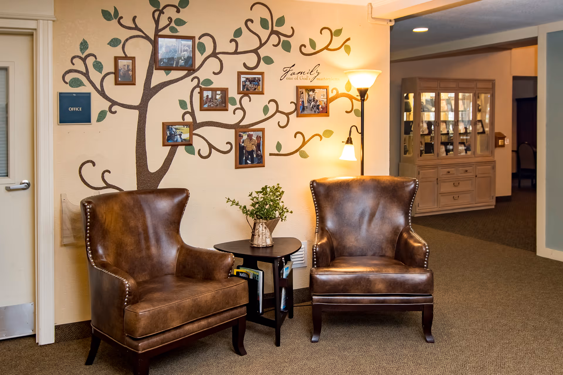 A cozy seating area with two brown leather armchairs and a small black side table with a plant on it. Behind the chairs is a wall decorated with a painted tree and several framed photos hanging on the branches. A floor lamp with two lights is positioned next to the chairs, providing warm lighting. To the left, there is a door labeled 'Office', and in the background, a wooden cabinet with glass doors is visible.