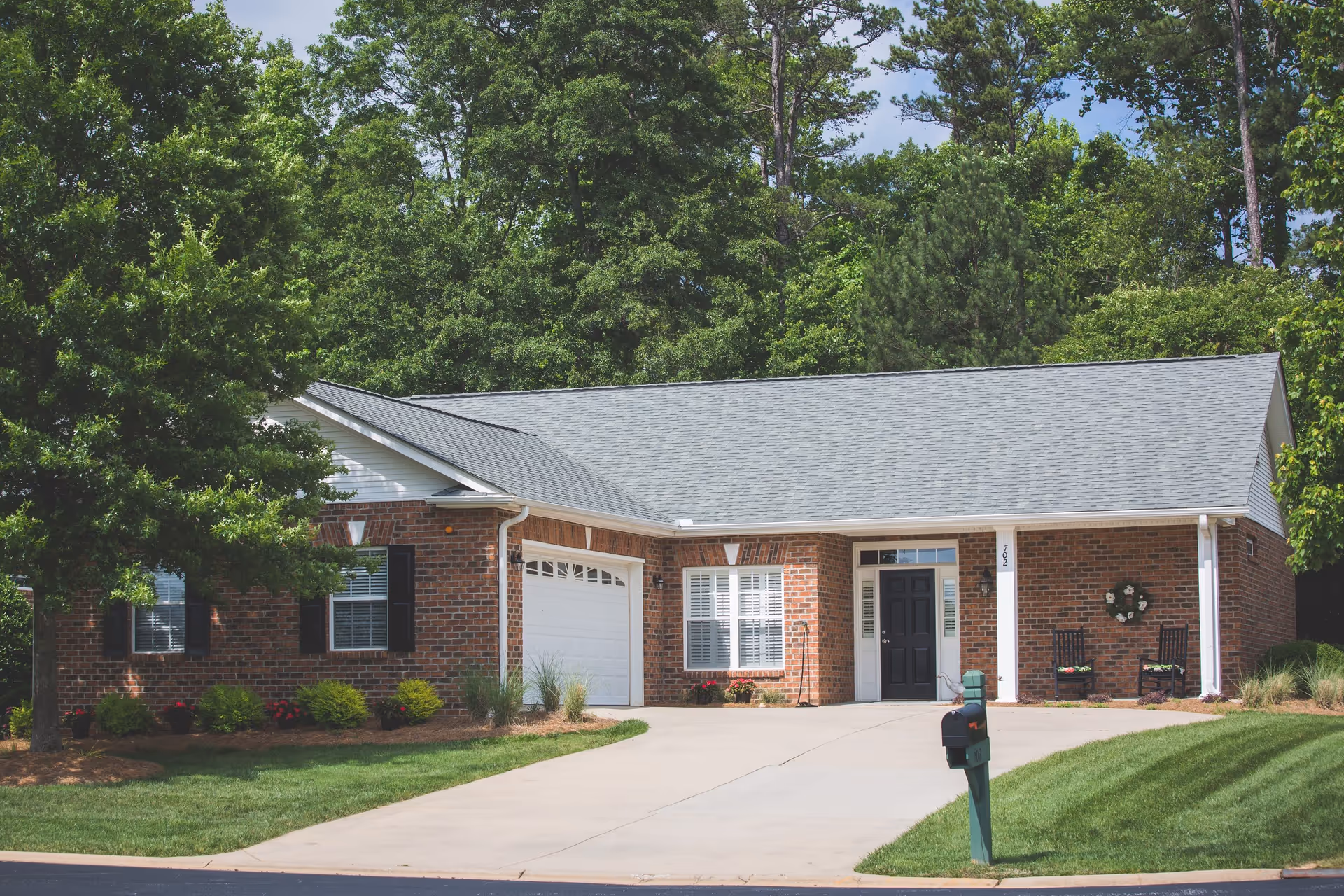 Single-story brick house with a gray shingled roof, white garage door, black front door, and two black rocking chairs on the front porch. The house is surrounded by green trees and a well-maintained lawn with a driveway and a mailbox in front.