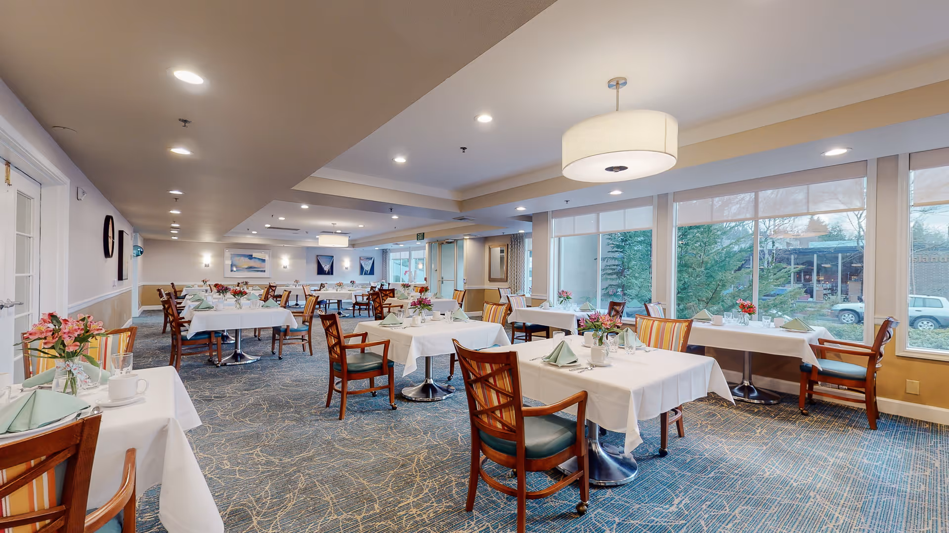 Bright, spacious dining room with multiple tables set with white tablecloths, chairs, and floral centerpieces beside large windows.