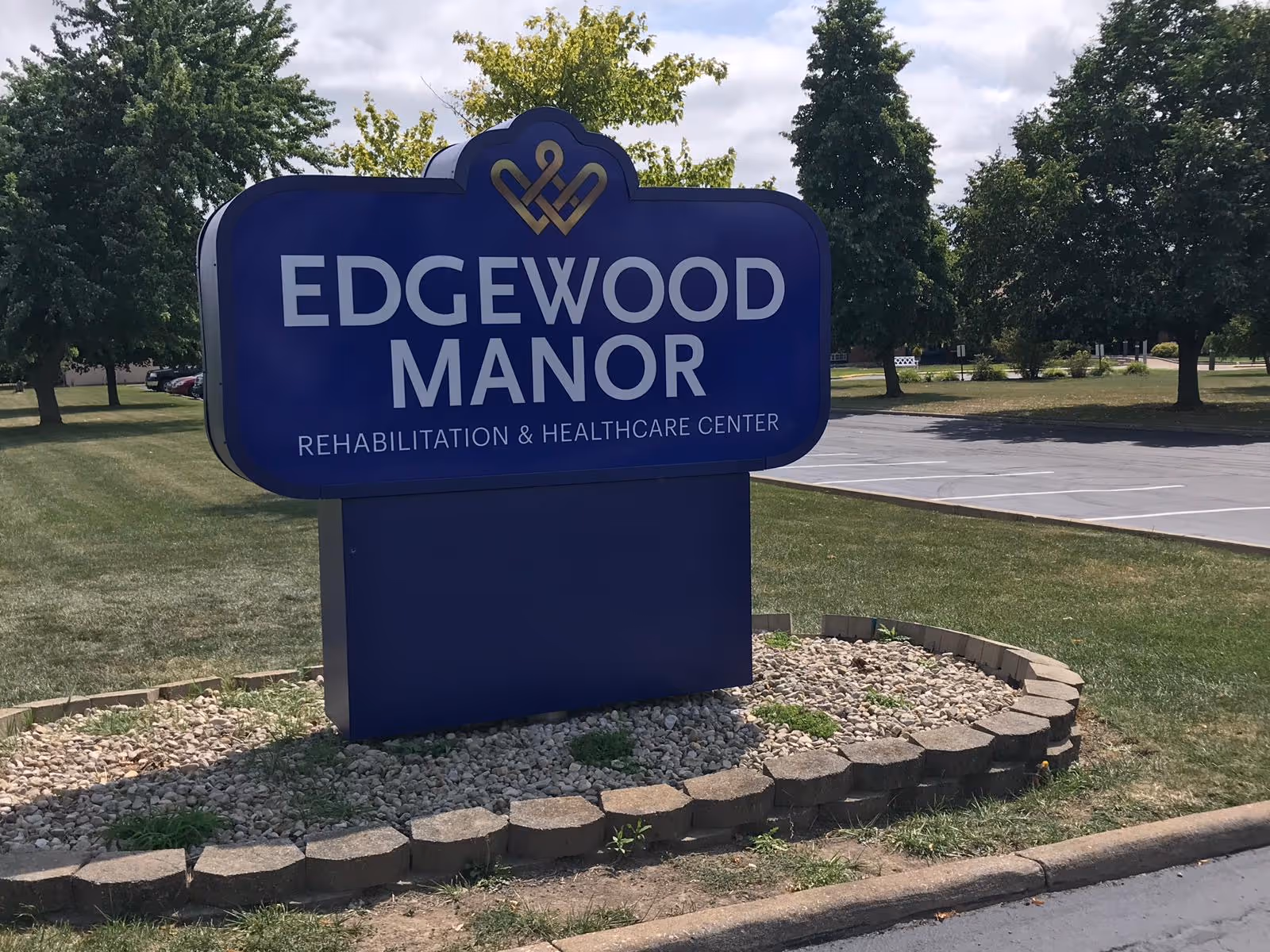 A large blue sign for Edgewood Manor Rehabilitation & Healthcare Center is displayed outdoors on a grassy area with trees and a parking lot in the background.