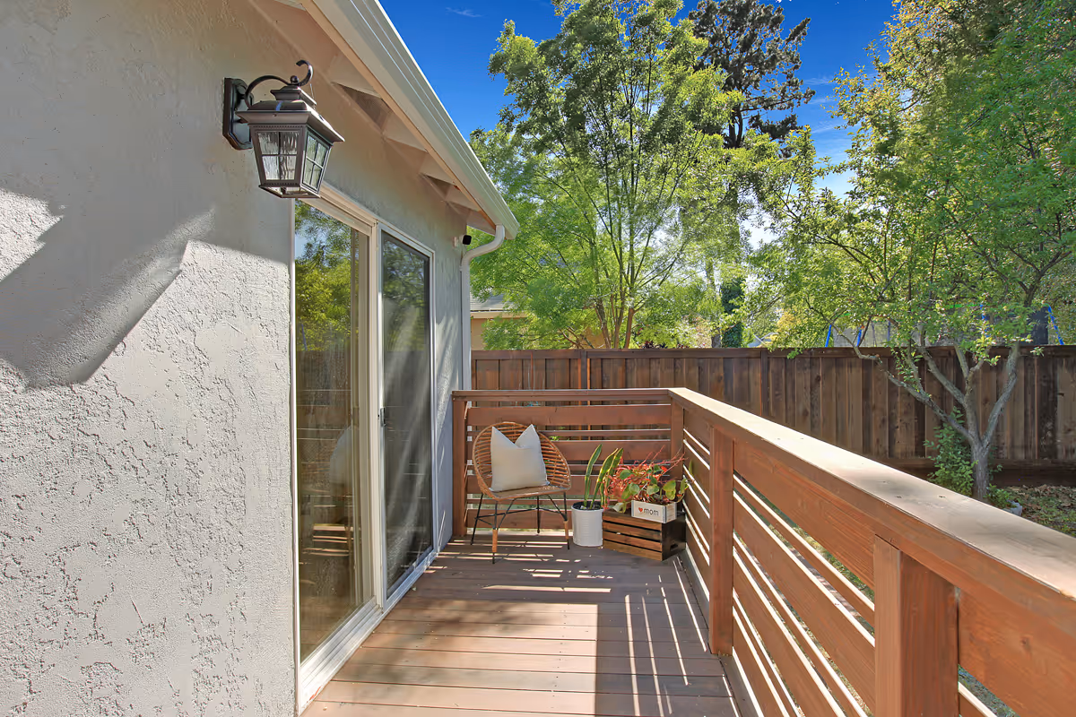 A wooden deck attached to a house with a sliding glass door. The deck has a wooden railing and a single chair with a cushion. There are two potted plants on the deck, and a wooden fence and green trees are visible in the background under a clear blue sky.