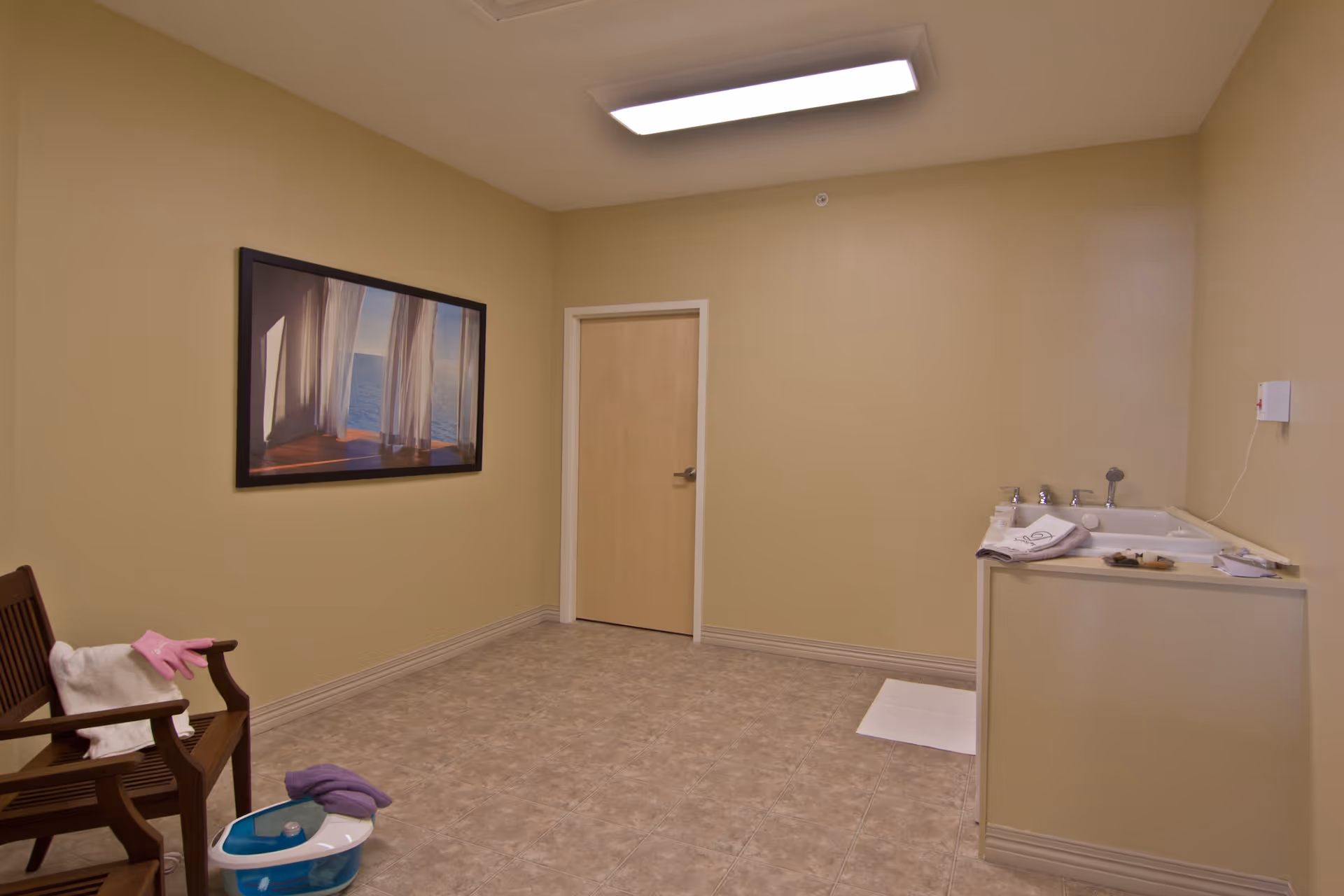 A small room with beige walls and tiled floor featuring a wooden bench with a white towel and pink gloves on it, a foot spa with a purple towel, a closed wooden door, a wall-mounted picture of a window with sheer curtains overlooking the ocean, and a small sink or basin area with towels and toiletries.