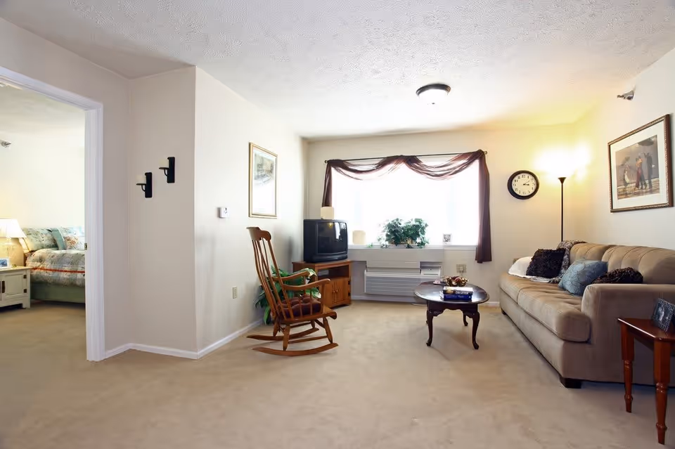 A cozy living room with a beige carpet and walls. There is a beige sofa with multiple cushions on the right side, a wooden rocking chair in the center, and a wooden coffee table with books and decorative items. A TV sits on a wooden stand near a large window with sheer brown curtains. A floor lamp and a wall clock are on the right wall, along with framed artwork. An open doorway on the left leads to a bedroom with a bed and nightstand visible.