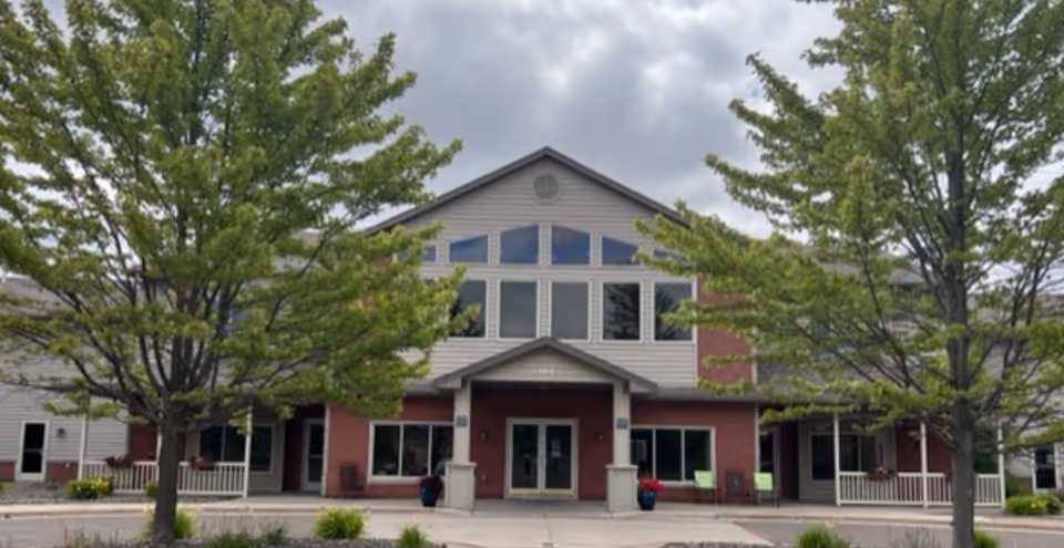 Front exterior view of Duluth Heights Lodge Senior Living building with a peaked roof, large windows, two trees in the foreground, and a covered entrance with double doors.