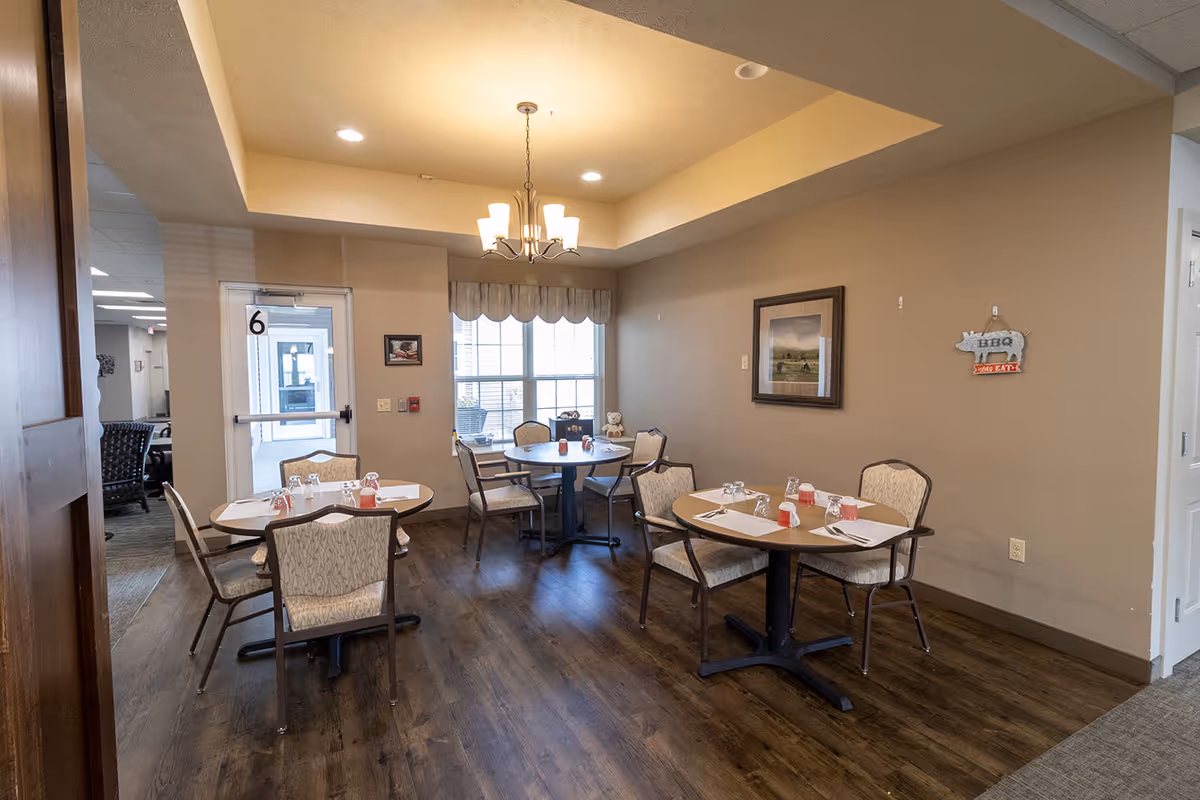 A small dining area in an assisted living facility with three round tables, each set with placemats, utensils, and glasses. The room has beige walls, a window with a valance, a ceiling light fixture, and framed artwork on the wall. There is a door with the number 6 on it leading to another area.