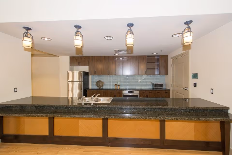 Modern kitchen area with a long dark granite countertop and sink in the foreground, wooden cabinets with a light blue tile backsplash, stainless steel refrigerator, oven, and a microwave on the right side. Four pendant lights hang from the ceiling above the counter.
