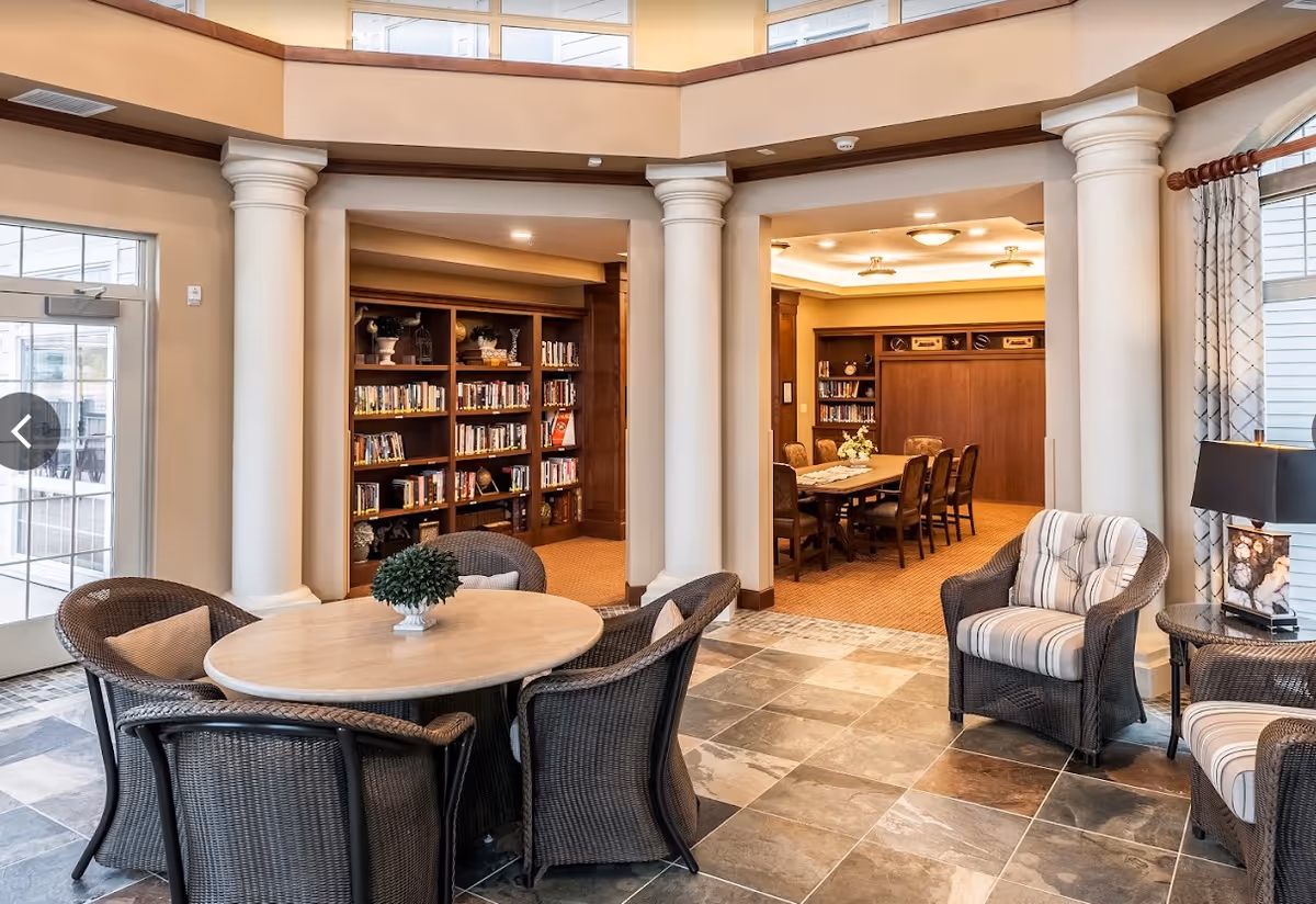 A cozy interior common area with a round table surrounded by wicker chairs with cushions, a small potted plant centerpiece, and two armchairs with striped cushions. The space features large white columns, tiled flooring, and a view into a room with a long wooden table and chairs, bookshelves filled with books, and warm lighting.